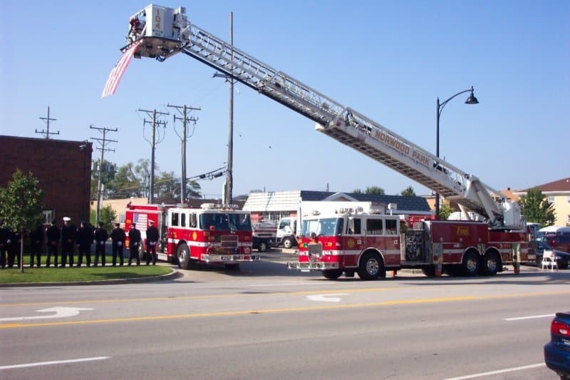 Firetrucks with extended ladder, American flag, firefighters lined up, street scene.