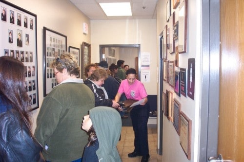People waiting in line in a hallway with wall photos and plaques, while a staff member in a pink shirt assists them.