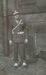 A person in vintage military or ceremonial uniform stands outdoors against a wooden building.