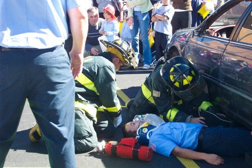 Firefighters are attending to a person lying on the ground next to a car, surrounded by onlookers.