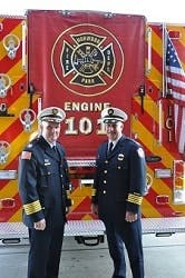 Two uniformed firefighters standing in front of a fire engine marked "Engine 101" with red and yellow chevron stripes and American flag.