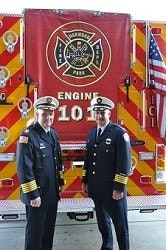 Two uniformed firefighters standing in front of a fire engine marked "Engine 101" with red and yellow chevron stripes and American flag.