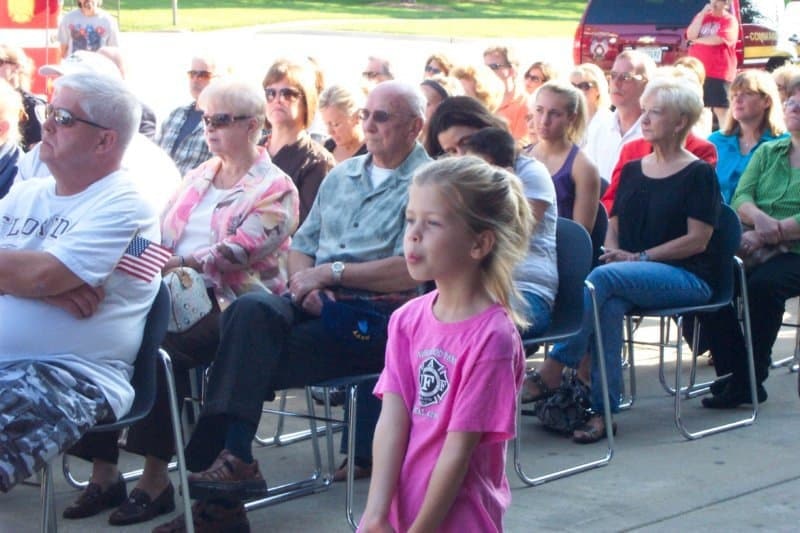 A young girl in a pink shirt stands in front of a seated crowd, some holding small American flags.