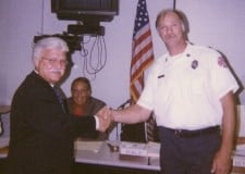 Two men shaking hands in an office setting, one in a suit and the other in a uniform, with an American flag in the background.