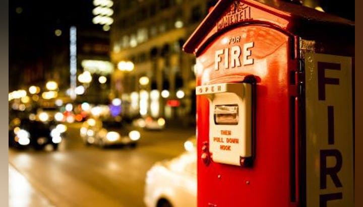 Red fire alarm box on a city street at night, with blurred lights and traffic in the background.