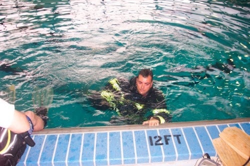 A person in a wetsuit is in a pool by the edge marked "12 FT."