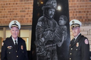 Two uniformed fire officials stand beside a monument depicting a firefighter carrying a child.
