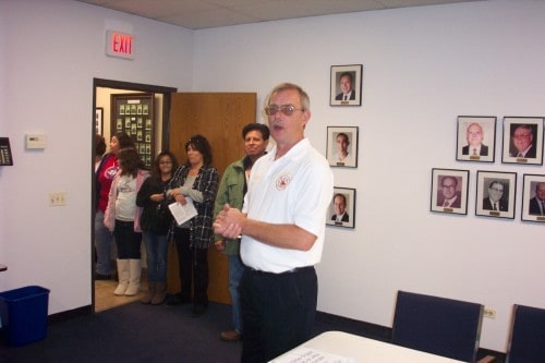 A person speaking to a group in a room with framed photos on the wall.
