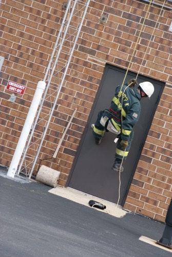 A person in safety gear rappelling down a brick building near a door.