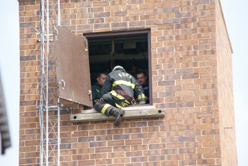 A firefighter climbs into a brick building through an open window, assisted by others inside.