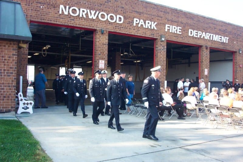 Firefighters in uniform march outside Norwood Park Fire Department; an audience is seated nearby.
