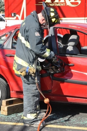 A firefighter uses hydraulic tools to open a damaged car door, with a fire truck in the background.