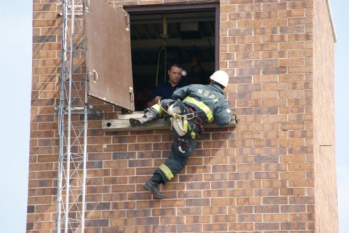 A firefighter is climbing into a brick building through an open window, assisted by another person inside.