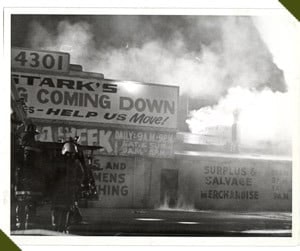 Firefighters battling heavy smoke at a burning building labeled "4301" with signs about moving and surplus merchandise.