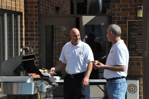 Two people in matching shirts are grilling hot dogs outside near a brick building.