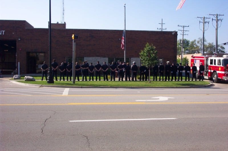A line of people in uniform stand solemnly in front of a building with a flag at half-mast, next to a fire truck.