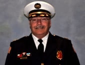 A person in a formal uniform and hat, featuring badges and insignia, against a neutral background.