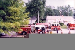 Fire trucks parked on a residential street with firefighters gathered nearby and trees in the background.