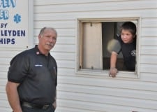A person stands outside a building while another person leans out of a window, both wearing dark shirts.