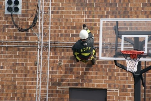 A firefighter in gear rappels down a brick wall near a basketball hoop.