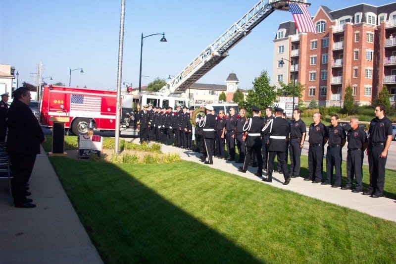 Firefighters in uniform gather in formation near a fire truck with an American flag, possibly for a memorial or ceremony.