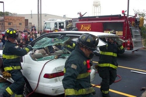 Firefighters demonstrate a car rescue, using tools to access a white vehicle, with a fire truck and crowd in the background.