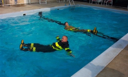 Two people floating in a pool wearing colorful firefighter turnout gear.