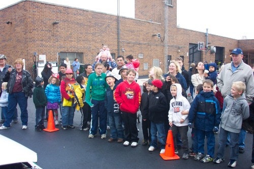 A group of people, including children, are gathered outdoors near orange cones, looking in the same direction.