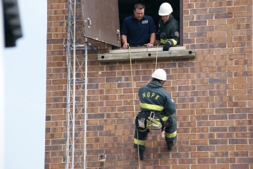 Firefighters conduct a rope rescue exercise, with one descending a brick wall while two others observe from a window.