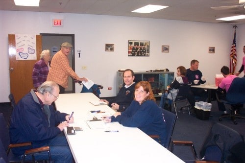 People sitting and standing around tables in a meeting room, with papers and an American flag in the corner.