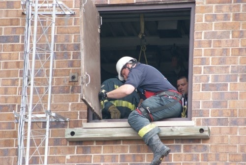 A firefighter in safety gear climbs through a window of a brick building.