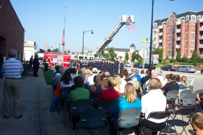 People are seated outdoors watching a ceremony with firefighters, a fire truck, and a raised flag on a ladder.