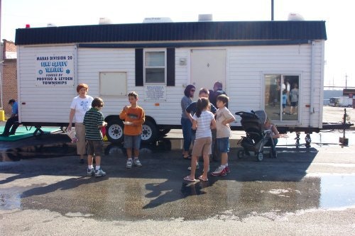 People stand near a mobile building with "MABAS Division 20" sign, on a sunny day. Children and adults are gathered outside.