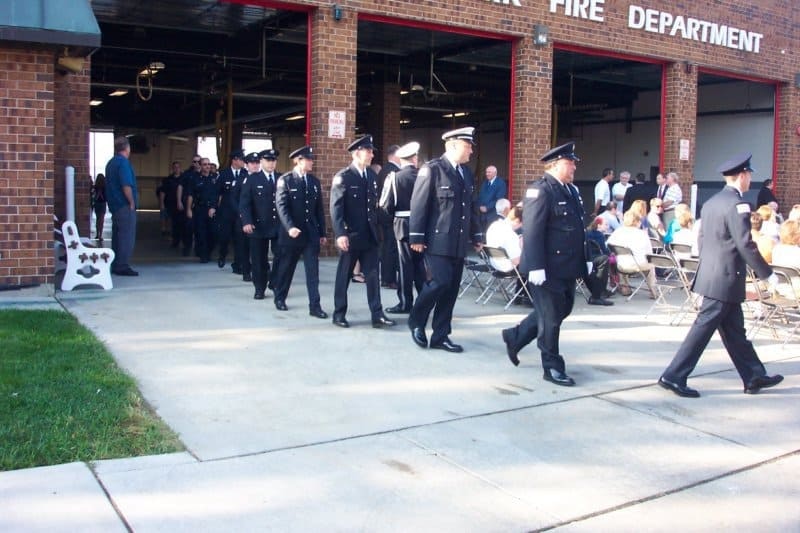 A procession of uniformed personnel walking past a crowd seated outside a fire department building.