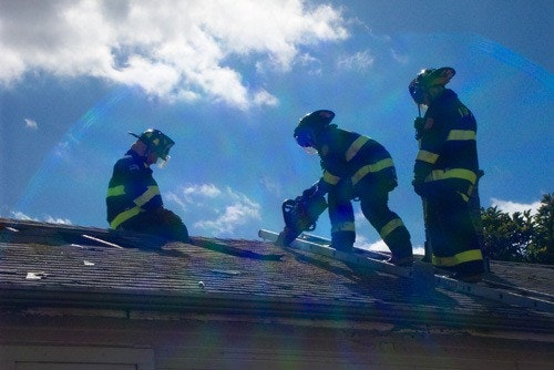 Firefighters in gear working on a rooftop under a sunny sky.