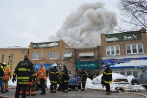Firefighters in action with smoke billowing from a building, surrounded by emergency personnel and vehicles.