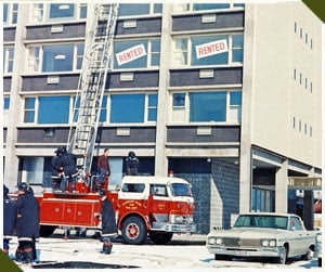 Fire truck with a ladder extended to a building marked "Rented," firefighters nearby, and a parked car in snowy surroundings.