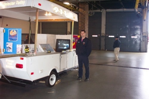 A person stands beside a mobile exhibit with a "911" sign. The setup includes informational displays and a monitor.