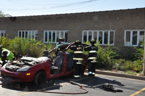 Firefighters in protective gear are using equipment to cut the roof off a red car in a parking lot.