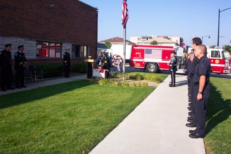 A memorial event with police and firefighters standing in formation near a fire truck and flag.