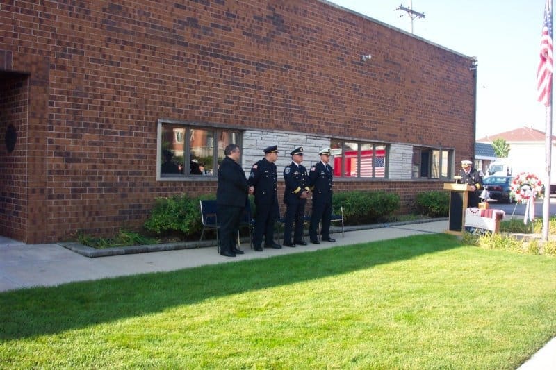 A group stands outside a brick building by a podium and flag. They appear to be in uniform, possibly at a ceremony.