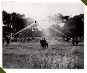 Two teams competing in a water hose challenge on a field, watched by a crowd, with water streams crossing in mid-air.