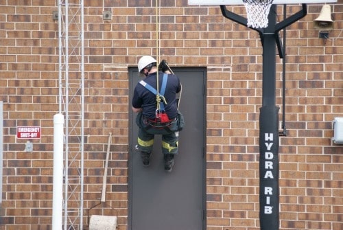 A person in safety gear and helmet climbs a brick wall using a harness near a door. A basketball hoop stands nearby.