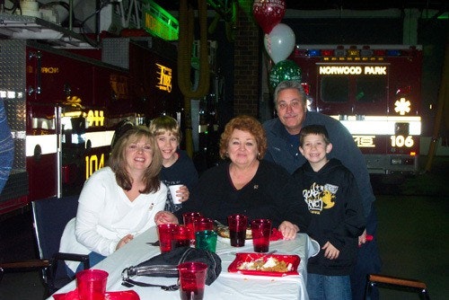 A group of people smiling at a table with food and drinks, with fire trucks in the background.