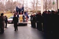 A ceremony with uniformed personnel and a fire truck carrying a casket draped in a flag, surrounded by people.