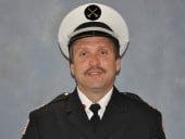 A man in a formal uniform with a white hat featuring a black emblem, against a plain background.