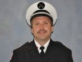 A man in a formal uniform with a white hat featuring a black emblem, against a plain background.