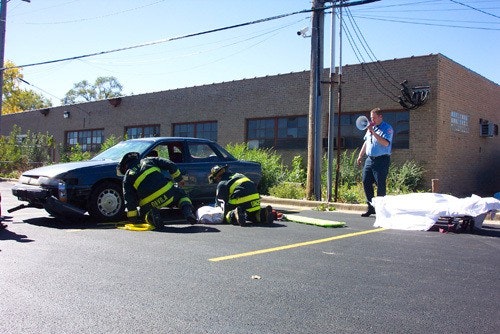 Firefighters are assisting a person near a car. Another person with a megaphone stands nearby. A covered figure is on the ground.