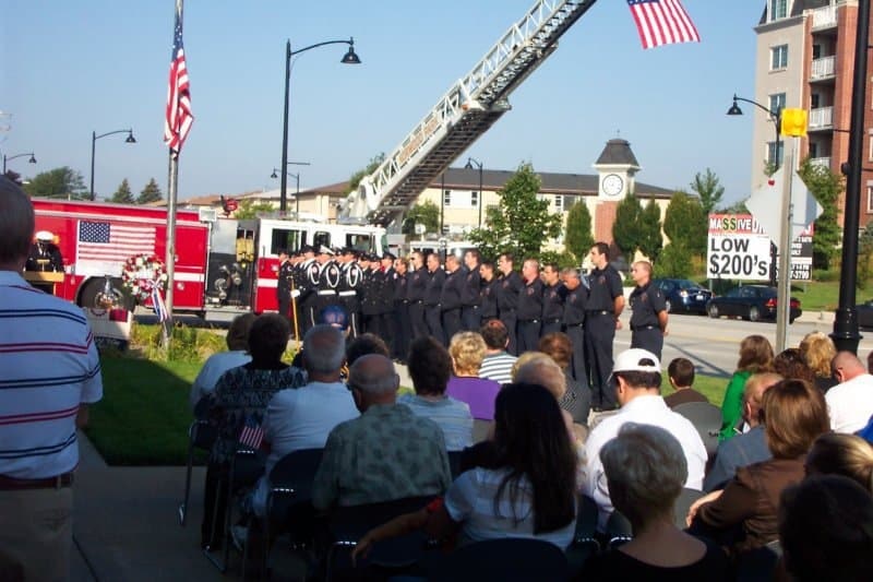 A gathering with firefighters in uniform, a fire truck, and American flags; people seated, observing the ceremony outdoors.
