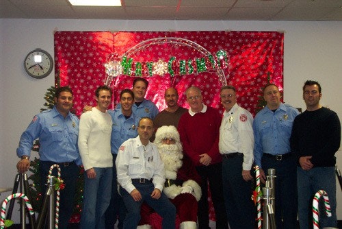 A group photo with people in blue uniforms and festive attire posing with Santa in front of a holiday-themed backdrop.